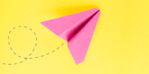 A pink paper airplane on a bright yellow background with a dashed line showing its flight path, illustrating how and why to post from Instagram to Pinterest for wider reach.