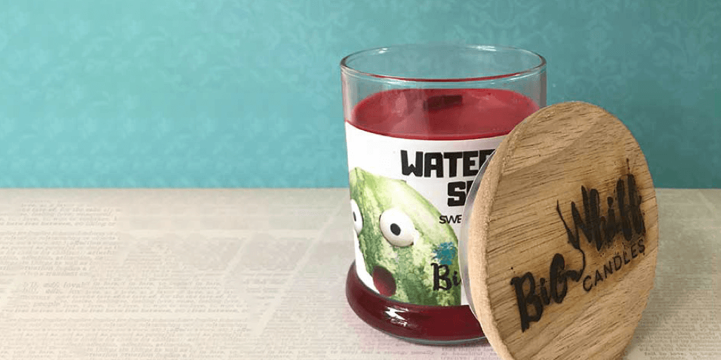 A red candle in a glass jar with a cartoon watermelon label stands on a surface; its wooden lid labeled “Big Wick Candles” leans against the jar, set before a teal patterned background—showcasing how to create gorgeous lifestyle photography.