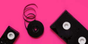Two black VHS tapes and a loose reel of unwound black tape are arranged on a bright pink background, reminding you that creating videos for Instagram has never been this easy.