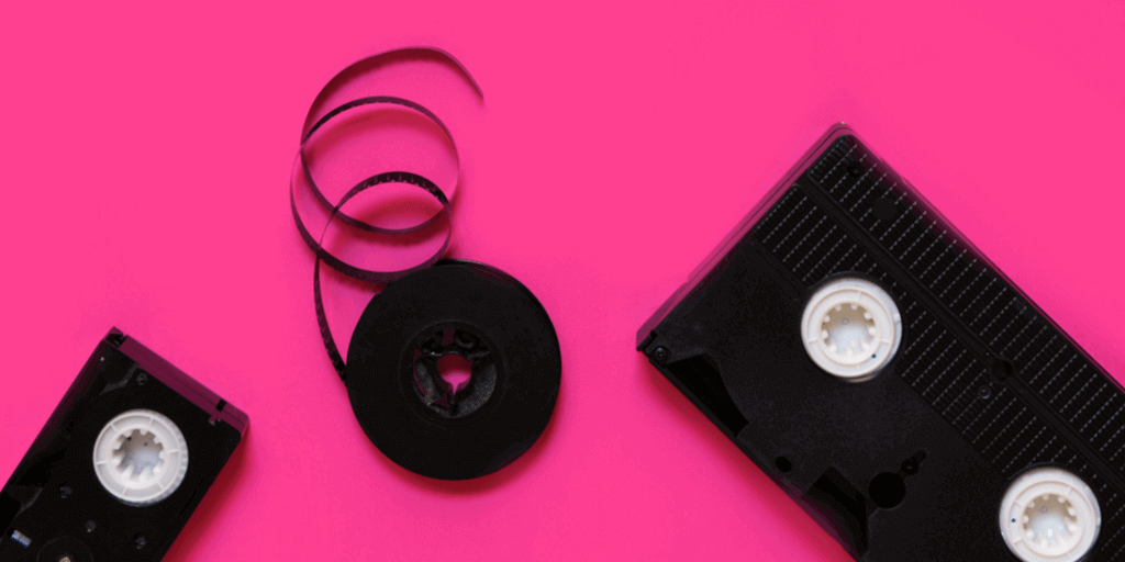 Two black VHS tapes and a loose reel of unwound black tape are arranged on a bright pink background, reminding you that creating videos for Instagram has never been this easy.