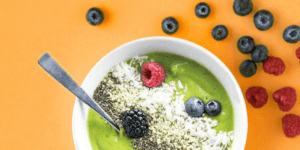 A bowl of green smoothie topped with shredded coconut, chia seeds, a blackberry, a raspberry, and blueberries sits with a spoon inside. More berries are scattered on an orange background—ideal for sharing photos or learning how to pin Etsy items on Pinterest.