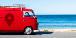 A bright red vintage van with a location pin symbol—like those used to show how to use geotags on Instagram posts and stories—is parked beside a sandy beach, with clear blue ocean waves and sky in the background.