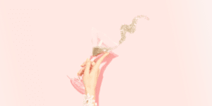 A hand holds a tilted martini glass with silver glitter spilling out in a swirl, set against a pastel pink background—perfect for showcasing how to find, save, and organize the best hashtags for Instagram.