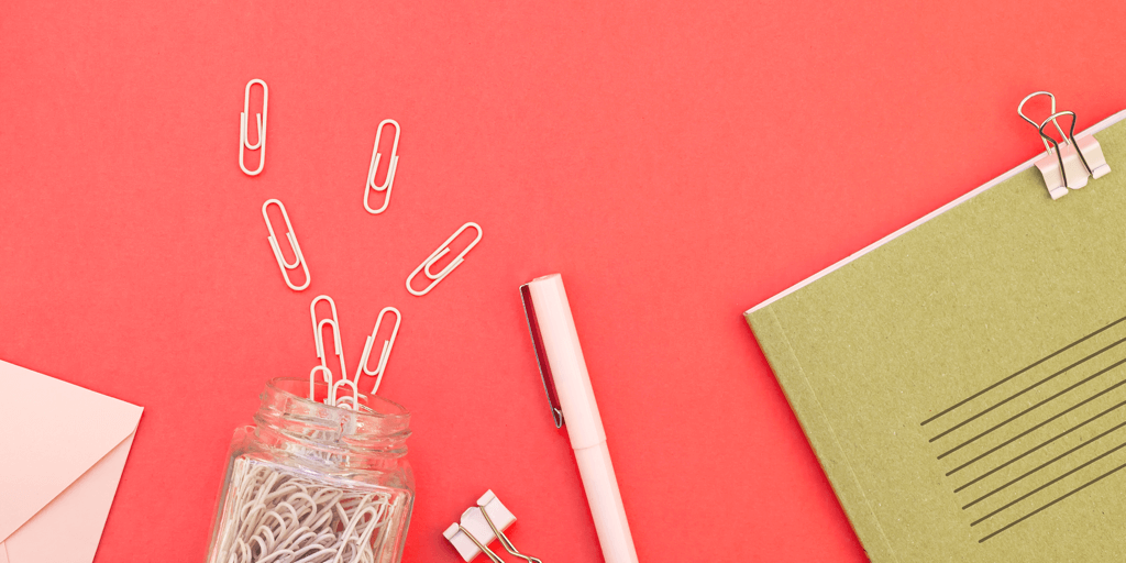 A jar of paperclips tipped over with paperclips spilling out, a pen, two binder clips, a green notebook, and a pink envelope on a red background.