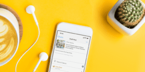 A smartphone displaying a social media draft post is on a yellow surface next to a cup of coffee, white earphones, and a small potted cactus, perfect for pondering questions like "should I auto post to Instagram?.
