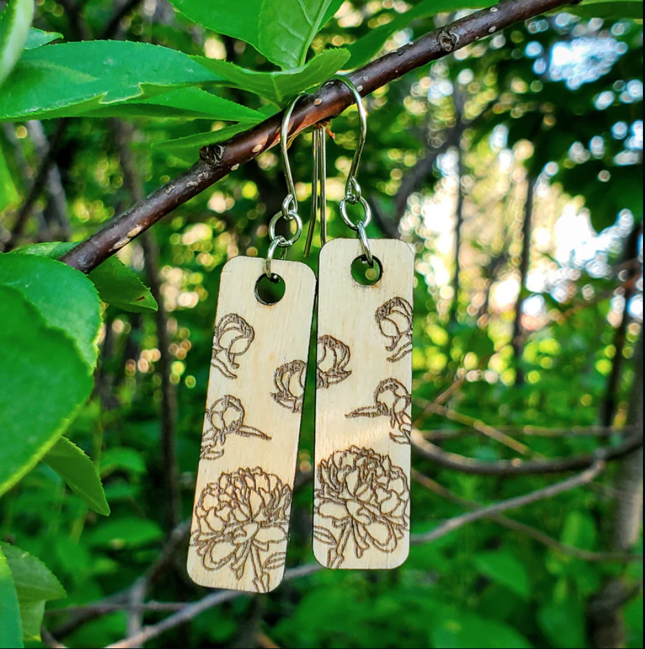 Screenshot of wooden earrings engraved with peonies hanging on a branch outside in the woods with green foliage behind.