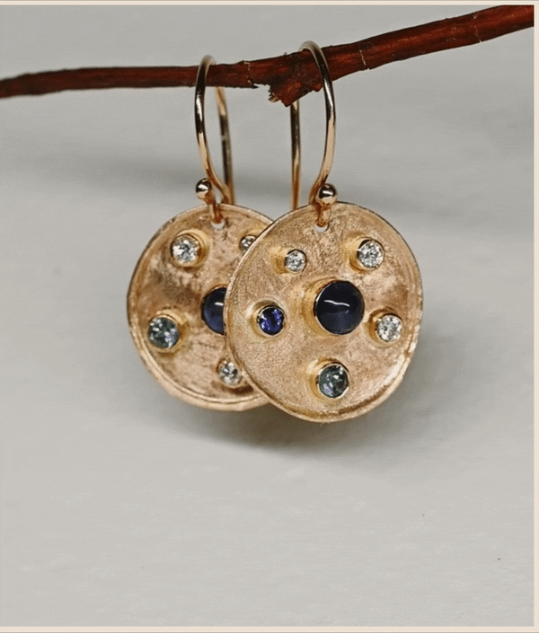 Screenshot of a gif of gold earrings with several inlaid stones hanging on a twig against a white background.