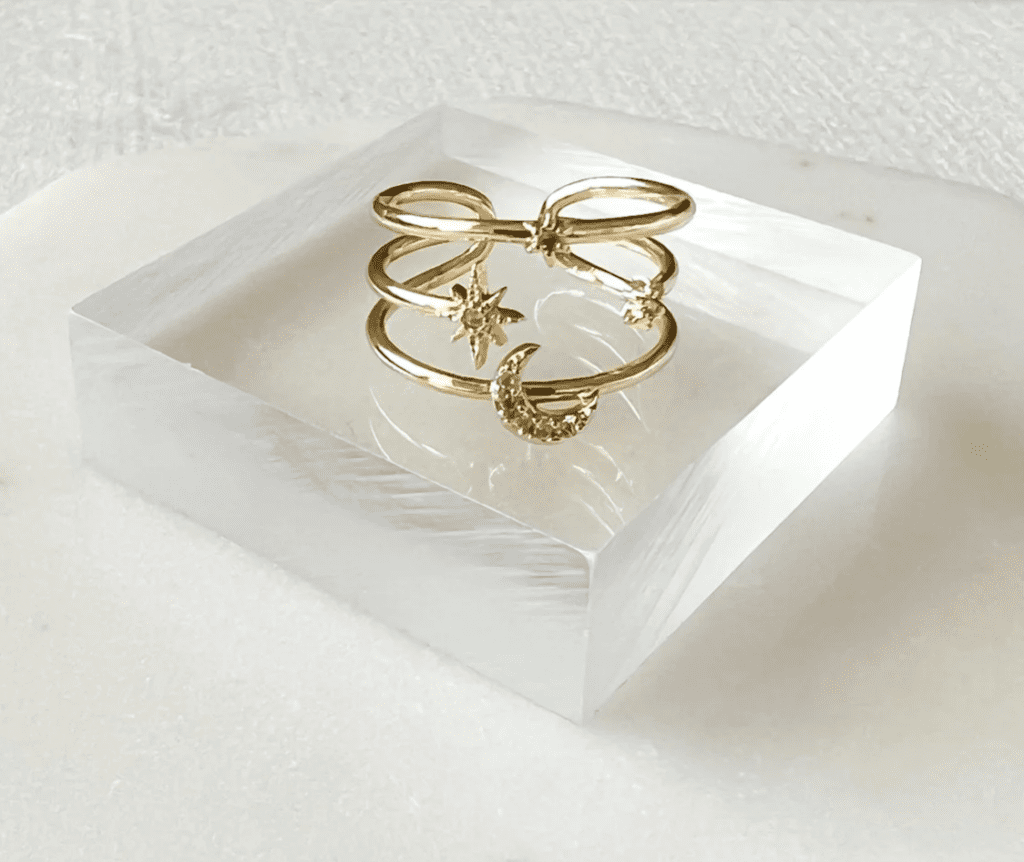Screenshot of a photo of a gold ring on a clear block stand on a white background.