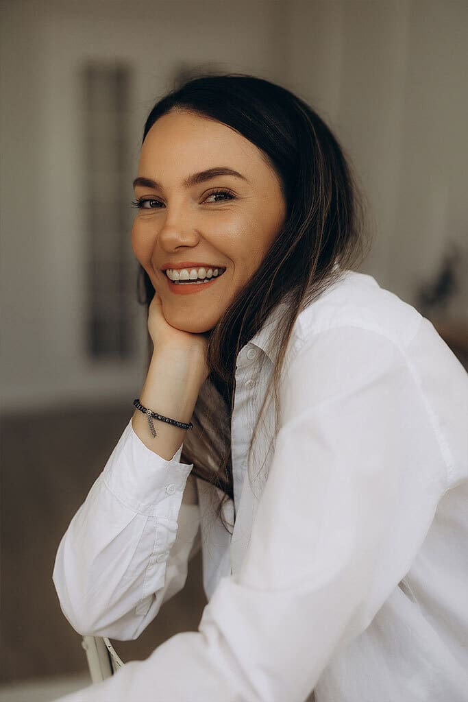 Photo of Yanna Burman, owner of urbanmamaz.com. She has long brown hair and is wearing a white long sleeved button down shirt and smiling at the camera.